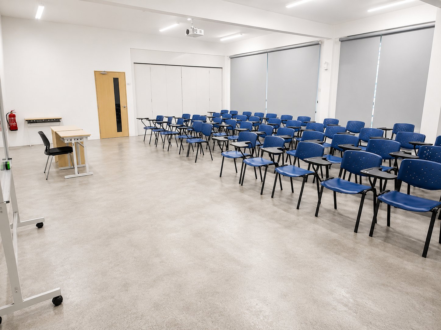 A refurbished classroom with rows of blue tablet-arm chairs, a lecturer's desk, a mobile whiteboard, and new roller blinds along the windows