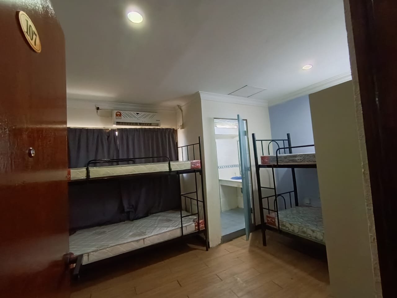 A hostel room with bunk beds either side of an ensuite bathroom doorway, an air conditioner, and privacy curtains