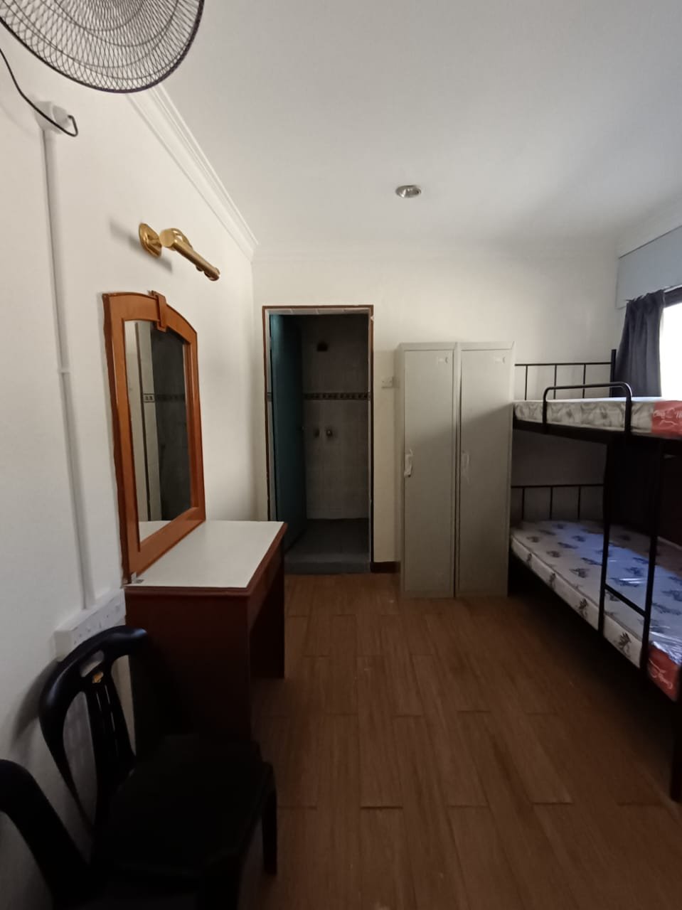 A hostel room with a dressing table and mirror, tall storage lockers, a wall fan, and a bunk bed with new mattresses