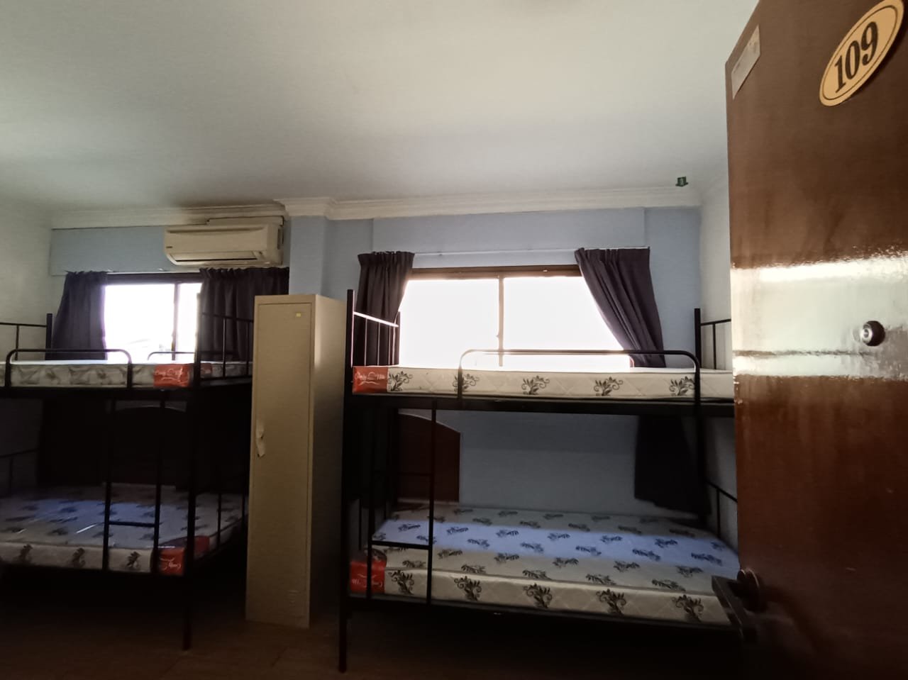 A hostel room with two sets of bunk beds and new mattresses, a storage locker, and curtained windows