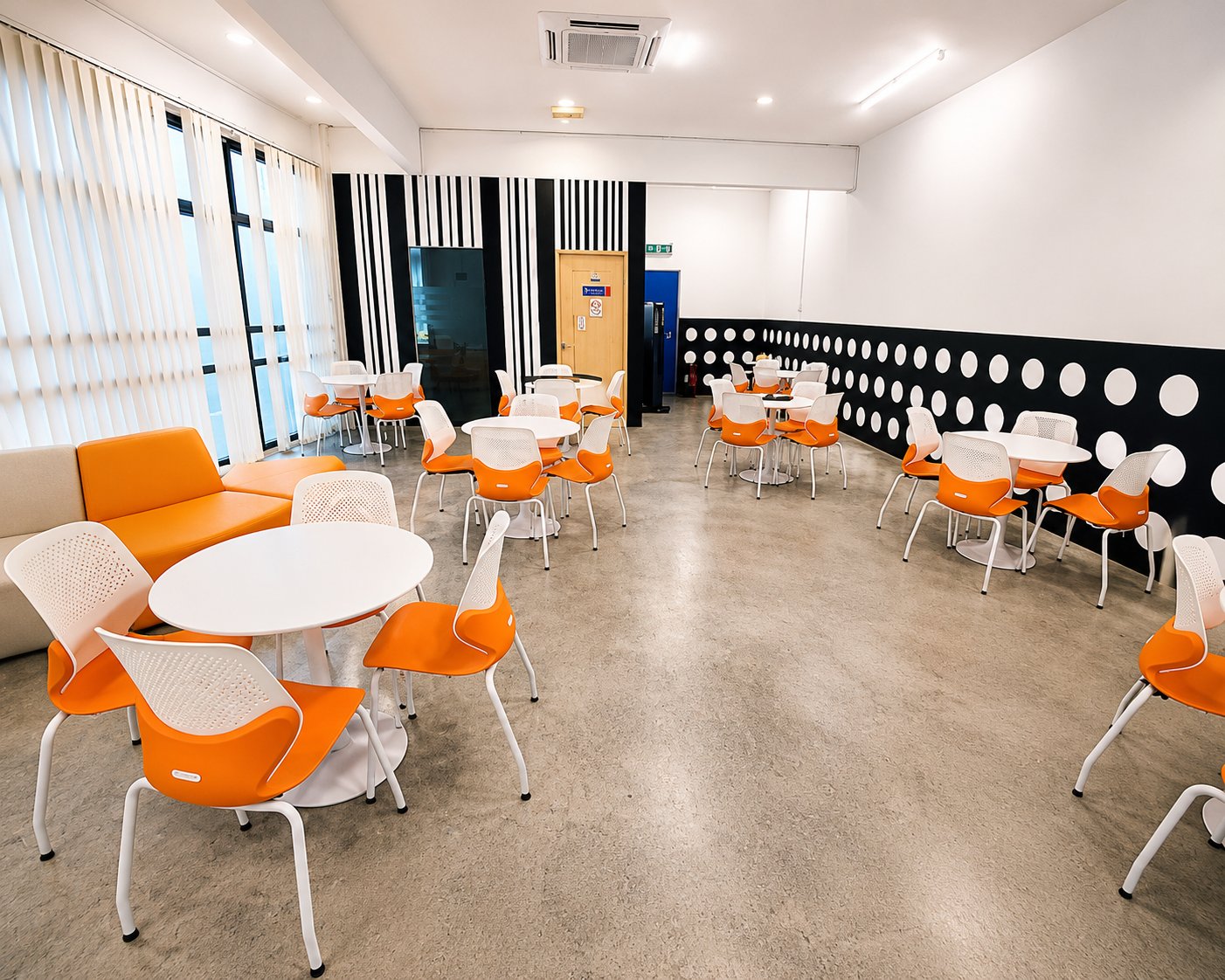 The refurbished student lounge, with round white tables, orange and white chairs, an orange banquette sofa, and a black feature wall with white circles