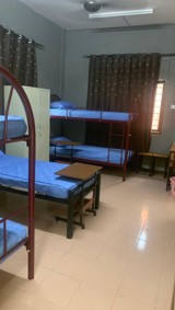 A hostel room with bunk beds, blue mattresses, bedside tables, and curtained windows