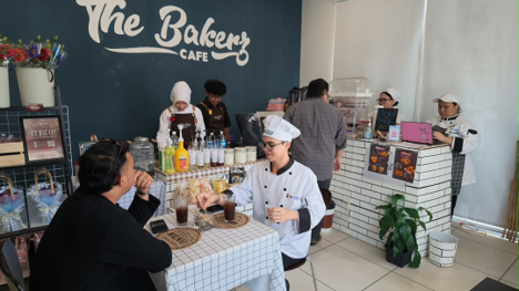 Students in chef whites serving customers at The Bakery Café counter, with a customer seated at a checkered-cloth table