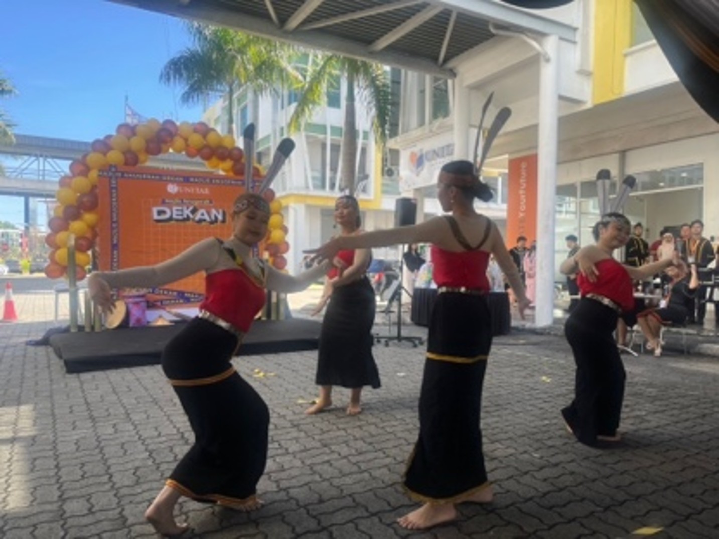 Students in traditional Sabahan dress performing a cultural dance outdoors, in front of a balloon arch reading Majlis Anugerah Dekan