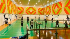 Players warming up on a badminton court in a hall with a brightly patterned mural wall
