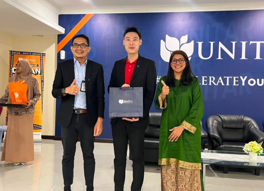 A guest speaker receiving a UNITAR gift bag from two staff members in front of the UNITAR #AccelerateYourFuture wall at an awards ceremony