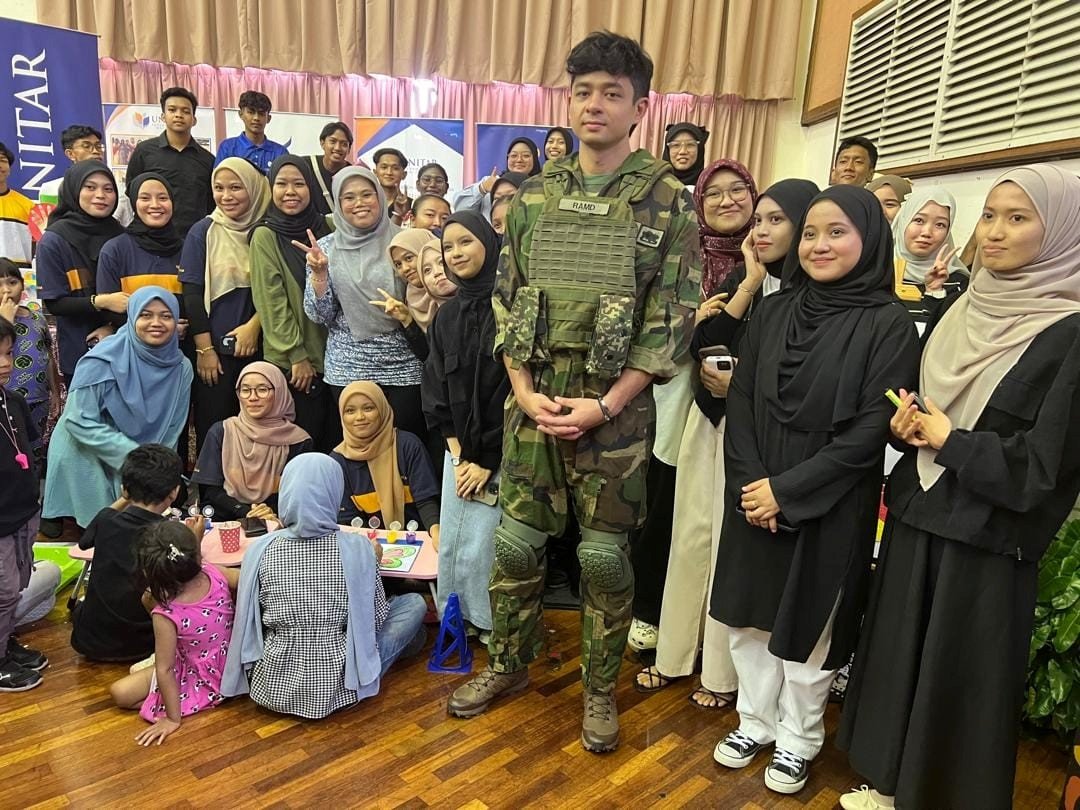 Students gathered around a serviceman in uniform at a community outreach event in a hall, with children colouring at a low table in the foreground