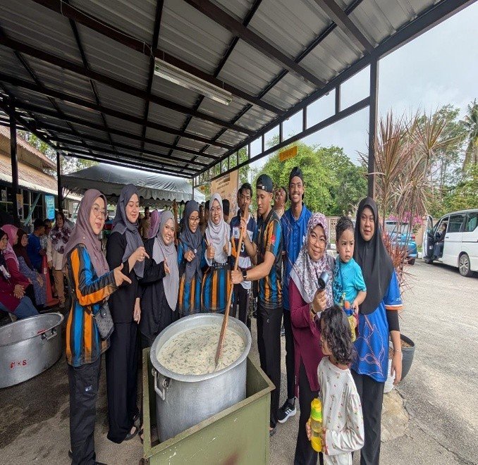 Students stirring a large cooking pot at an outdoor community kitchen, alongside local residents and children