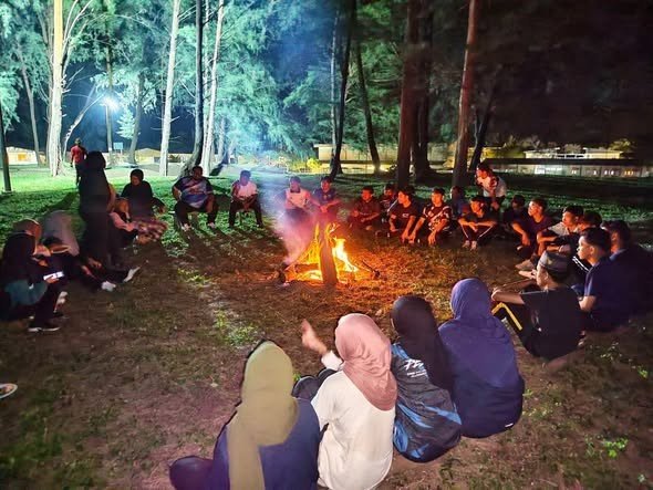 Students sitting in a circle around a campfire among trees at night during a camp