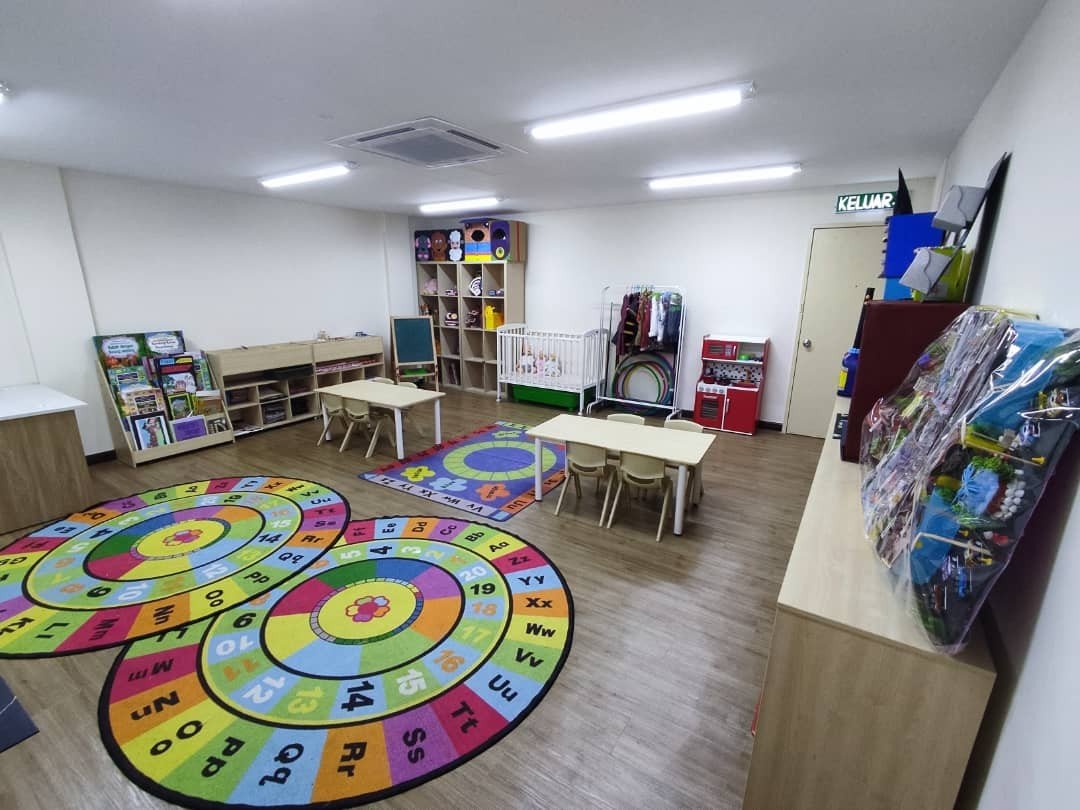 A mock kindergarten teaching room with alphabet and number rugs, child-sized tables and chairs, a book display, a play kitchen, and a cot