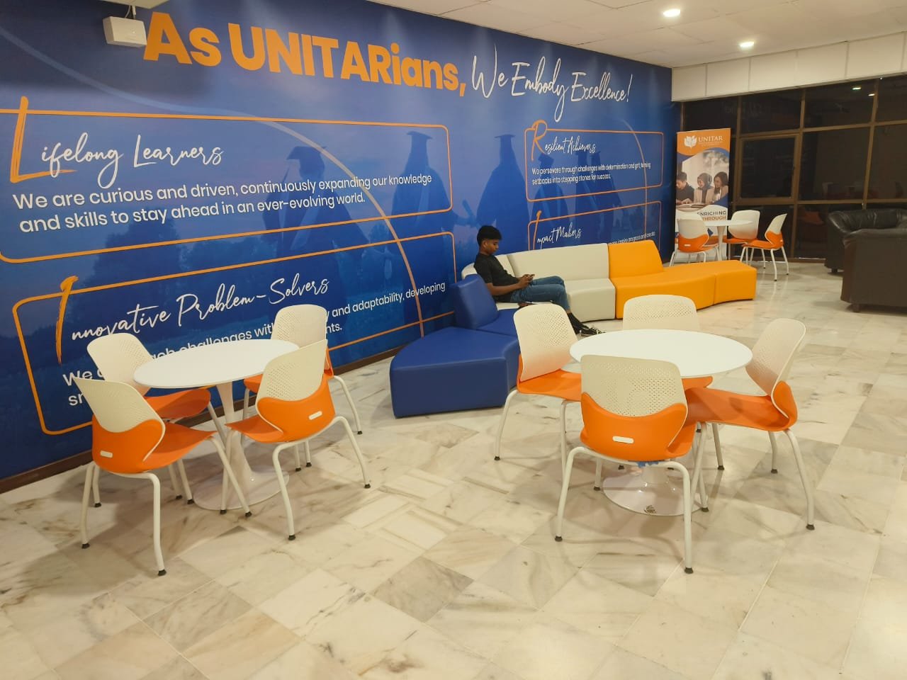 A common area with round tables, orange chairs, and blue and orange modular seating against a mural reading 'As UNITARians, We Embody Excellence'
