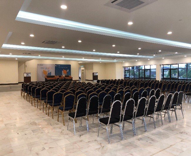 A large hall laid out with rows of banquet chairs facing a stage, with windows along one side