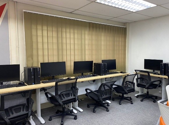 A second computer lab, with new monitors and desktop towers along a bench beneath vertical window blinds