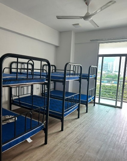 A hostel room with new blue-mattressed bunk beds, a ceiling fan, and a balcony looking out over greenery