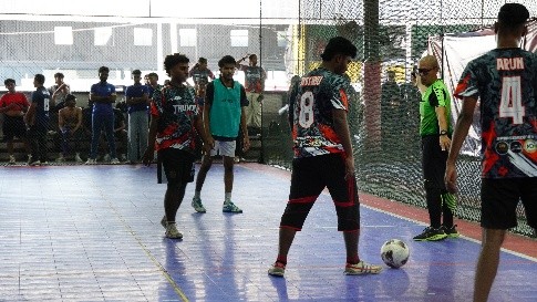 Students playing futsal on an indoor court while teammates watch from the sideline
