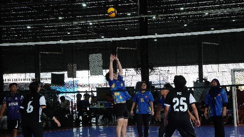 Students reaching for the ball during a volleyball match, with spectators watching from the side