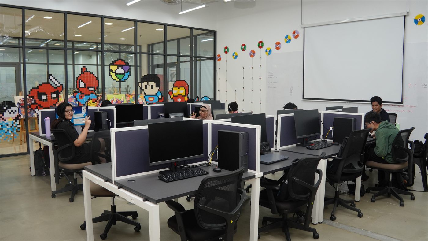 Rows of desktop workstations in a computer lab, with students working at the machines and pixel-art murals on the glass partition behind