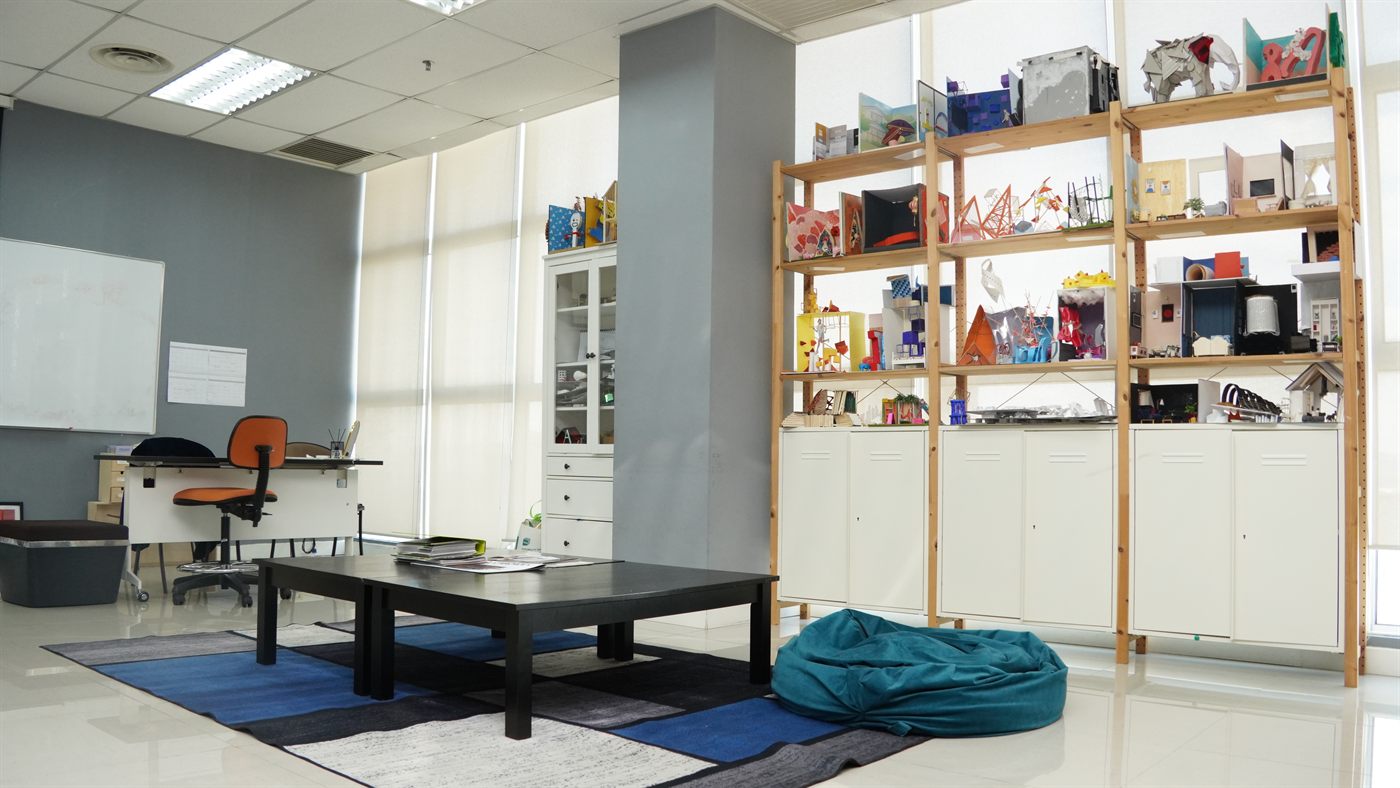Open shelving filled with student-built scale models, beside a low table, rug, and bean bag in a studio space