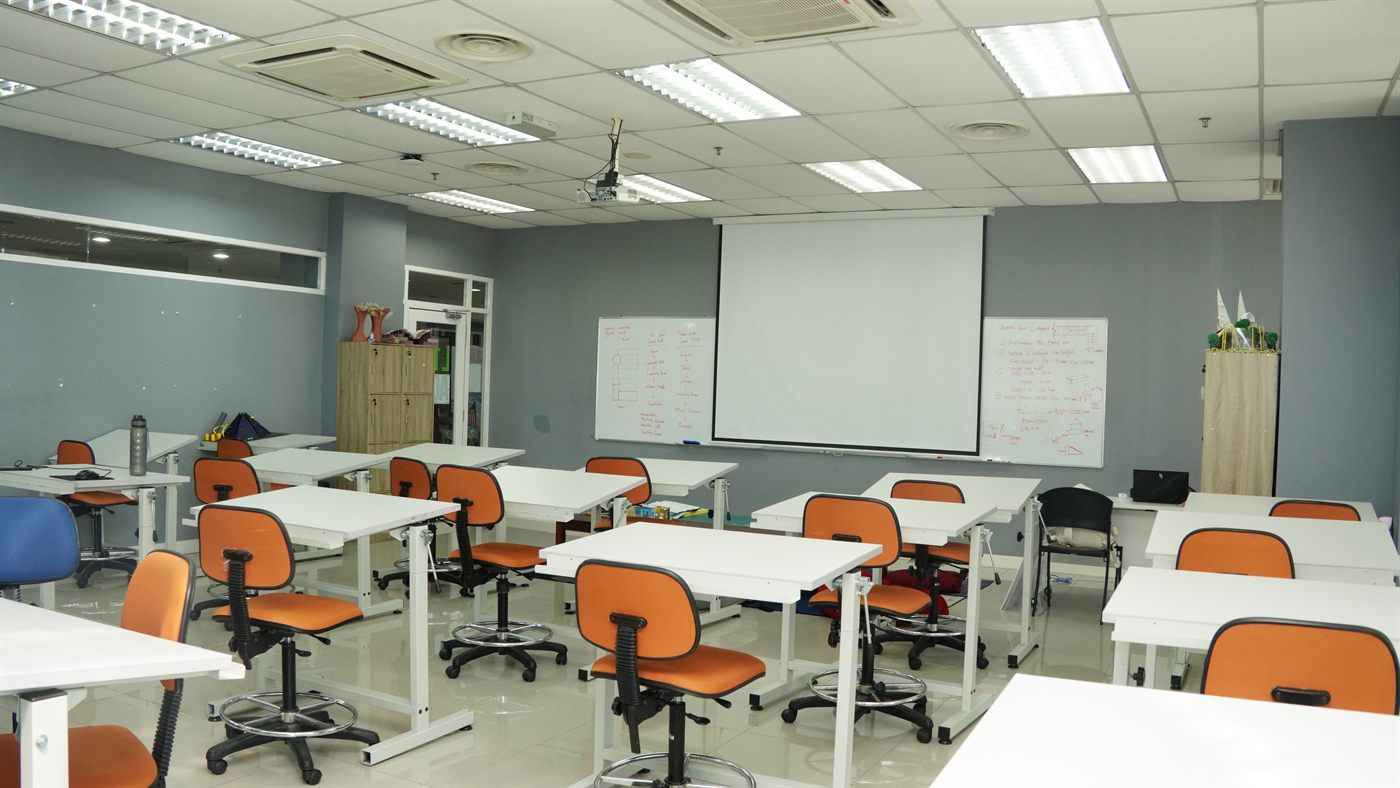 A studio classroom filled with adjustable drafting tables and orange drafting stools, facing a whiteboard and projector screen