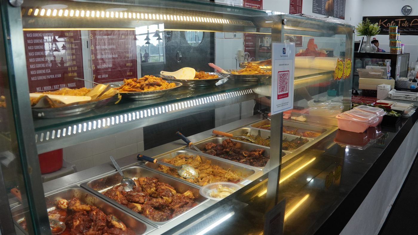 The café counter, with cooked dishes in the display case and a menu board listing prices from RM7 to RM10