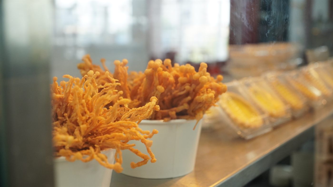 Cups of crispy fried enoki mushrooms on the counter, with egg tarts lined up behind