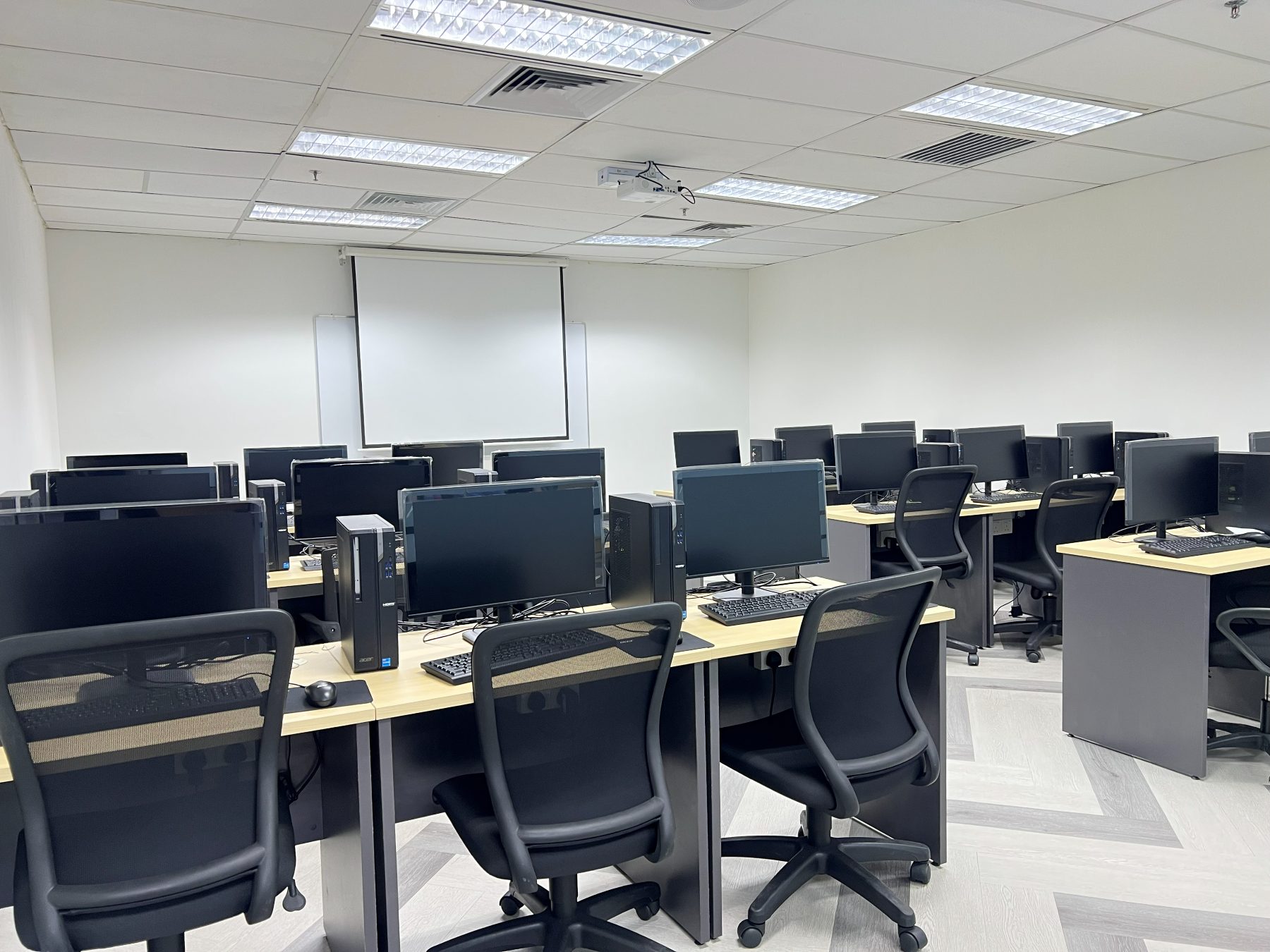 A computer lab with rows of desktop workstations facing a whiteboard and projector screen, with a projector mounted on the ceiling
