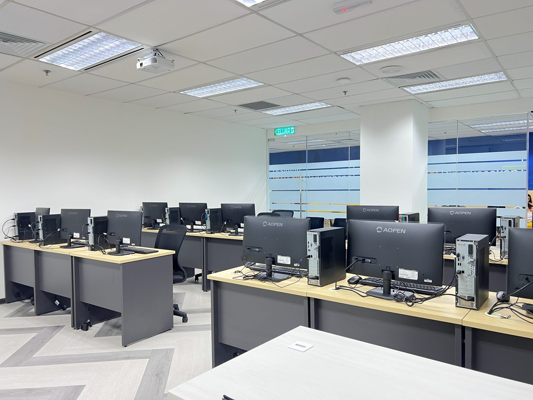 A computer lab with rows of monitors and desktop towers on light wood benches, lit by ceiling panels