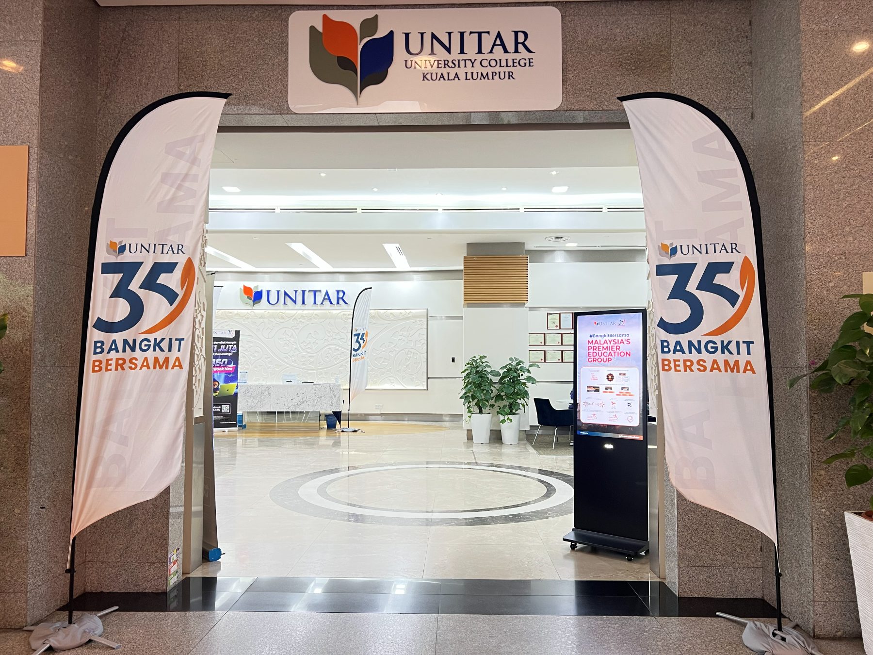 The UNITAR University College Kuala Lumpur entrance off the building lobby, with UNITAR 35 Bangkit Bersama flags either side of the doorway and the reception counter beyond
