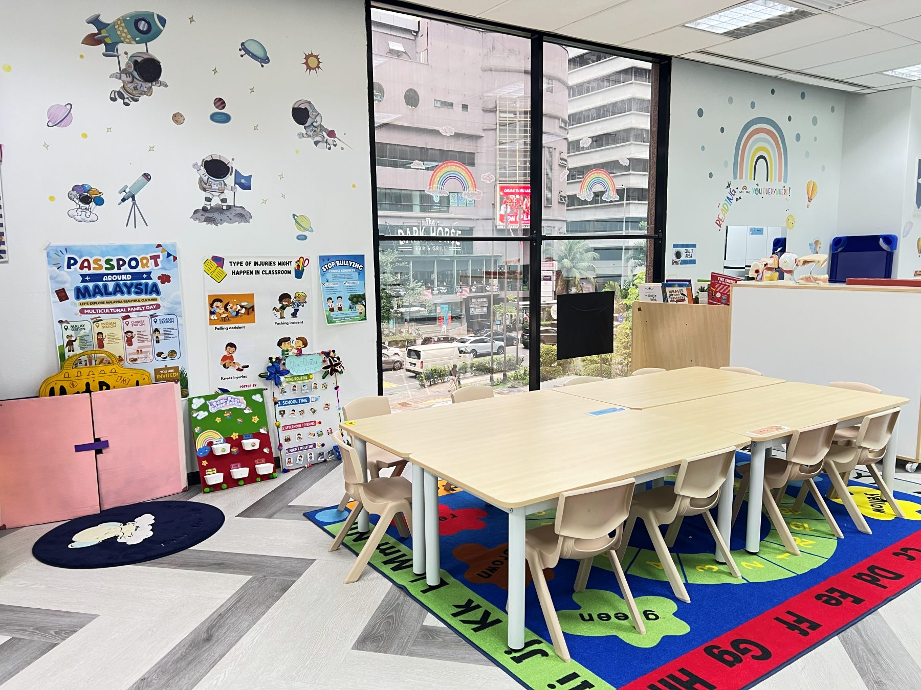 A mock kindergarten classroom with a low table and child-sized chairs on an alphabet rug, teaching posters on the wall, and a window looking onto the street