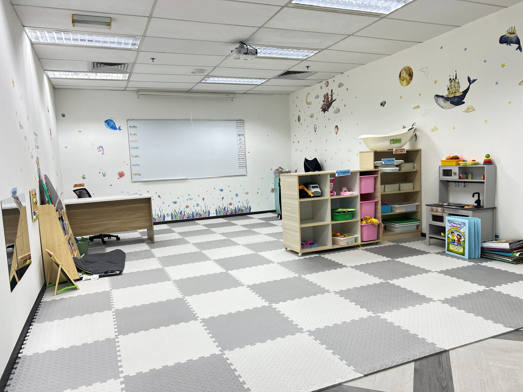 A mock kindergarten room with foam floor mats, a whiteboard carrying a weekly routine chart, toy storage shelving, and a play kitchen
