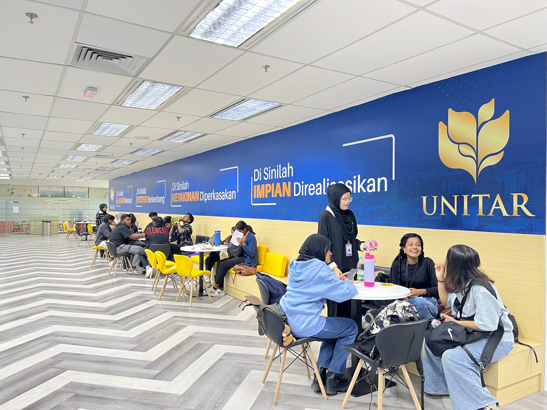 Students sitting at round tables and yellow chairs in the Level 2 student lounge, along a long blue UNITAR feature wall