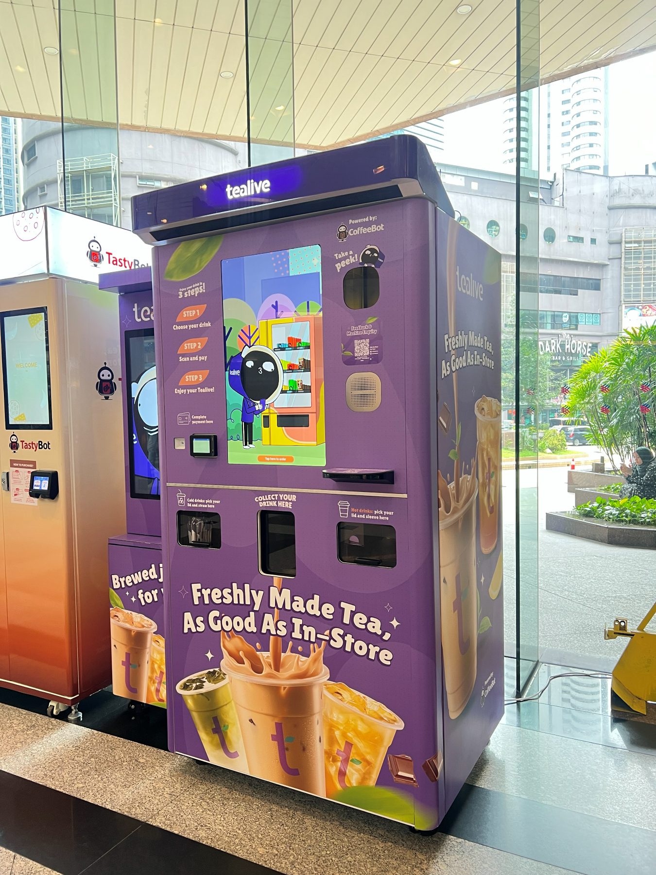 A purple Tealive vending machine beside the TastyBot machine, with a touchscreen showing a three-step ordering guide
